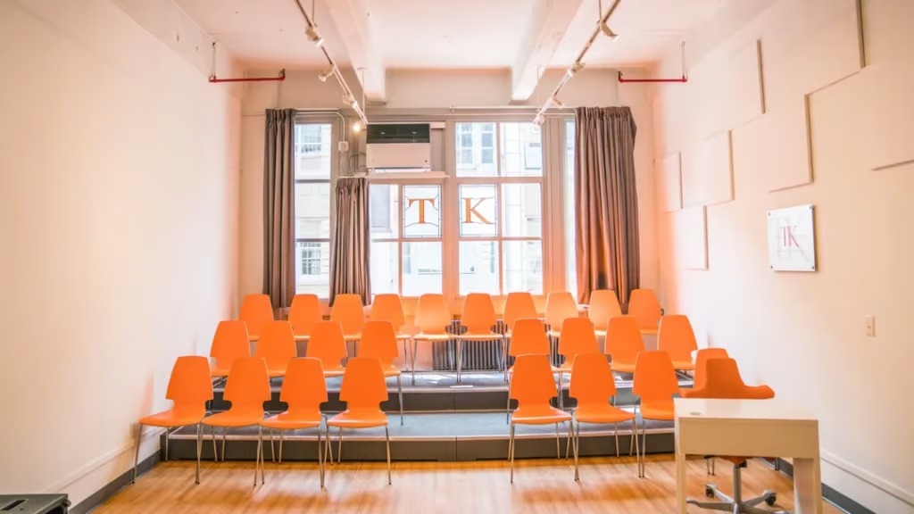 A minimalist classroom with several rows of bright orange chairs facing a desk at the front. Large windows in the back let in natural light, and walls are adorned with acoustic panels and curtains. A sign with "TK" is visible, while a compact module library sits unobtrusively in the corner.