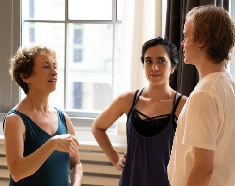 Three people stand in a brightly lit room near a window. A woman gestures while speaking to a man and another woman, who are both listening attentively. They appear to be having a discussion about the new module library in a casual setting.