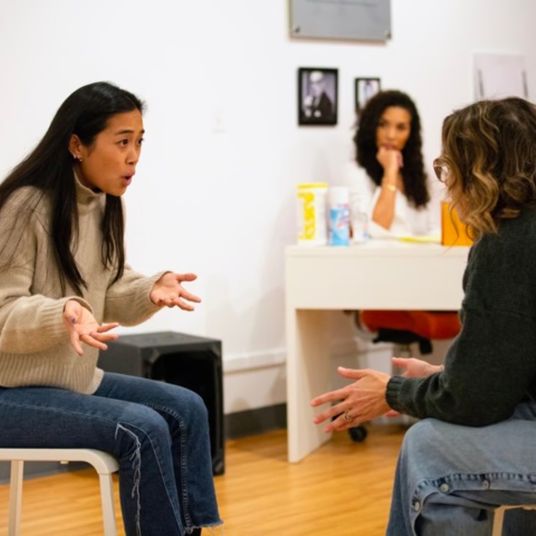 Two women are sitting on chairs in deep conversation, with one gesturing expressively. In the background, another woman at a desk observes them, surrounded by photos and a sanitizer. The cozy room, resembling a module library, features a wooden floor and pristine white walls.