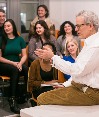 During an Open House, a group of students attentively listens to a man in a white shirt, seated on a desk in the classroom. Casually dressed, they take notes while sitting on orange chairs arranged in a semicircle.