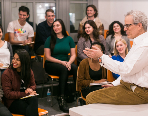 During an Open House, a group of students attentively listens to a man in a white shirt, seated on a desk in the classroom. Casually dressed, they take notes while sitting on orange chairs arranged in a semicircle.