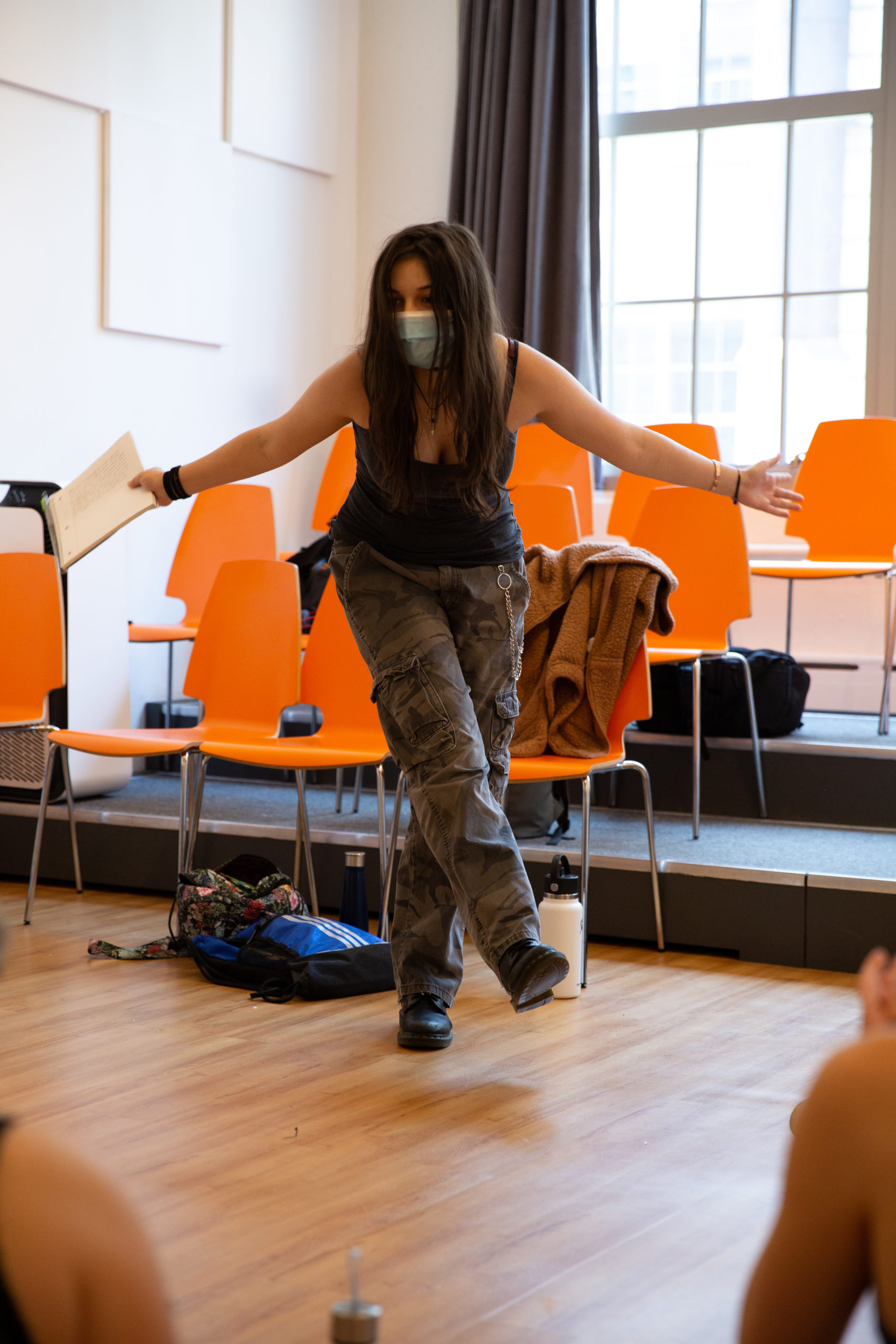 A woman wearing a mask and casual clothing, part of the Summer High School Faculty, holds a notebook while balancing on one foot inside a room. Bright orange chairs and personal items lend a celebratory touch to the scene.