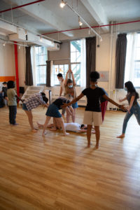 A group of people is standing in a circle, holding hands, in a brightly lit room with wooden floors and large windows. One person, perhaps part of the Summer High School faculty, lies at the center. Everyone appears to be engaged in an activity.