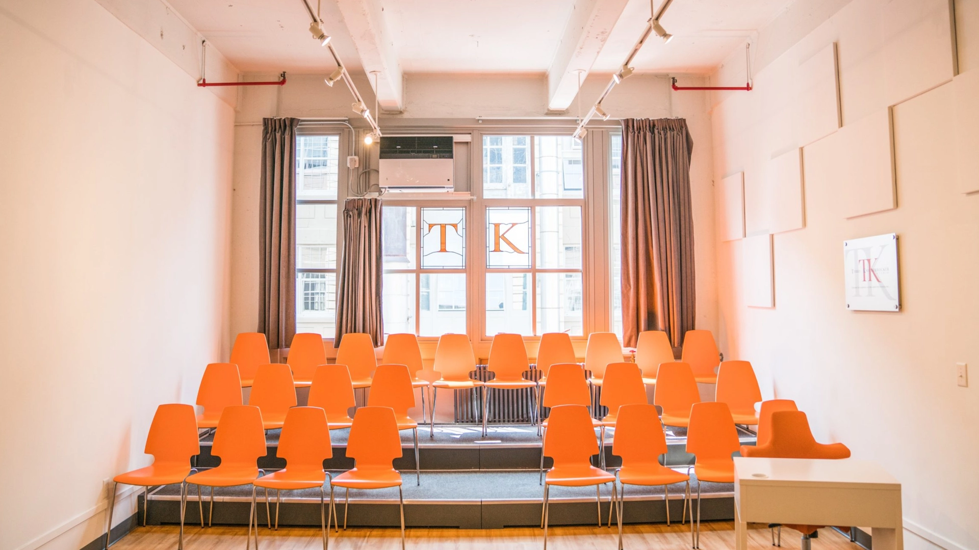 A bright, airy room designed for actor training features rows of orange chairs facing a small stage. Large windows at the back let in natural light, complemented by white sound panels on the walls and a framed logo on the right wall. Curtains elegantly frame the windows.