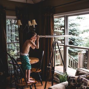 A young boy in swim shorts stands barefoot on a chair indoors, embodying a beginner's mindset as he eagerly peers through a telescope pointed out a large window. The room, with its wooden furniture and dark curtains, offers him an expansive view of the trees outside.