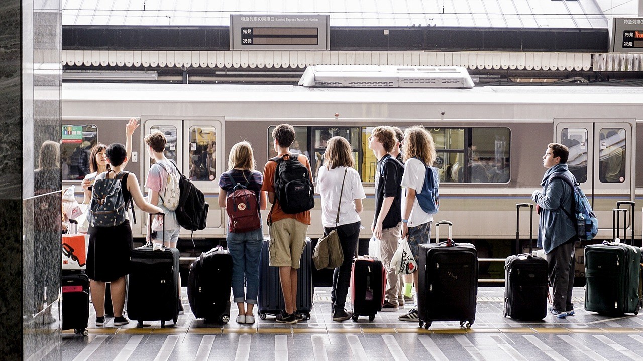 A group of people with backpacks and luggage stand on a train platform, facing a stationary train. They appear to be waiting or preparing to board, perhaps seeking inspiration for their journey. The covered platform features signs with text visible above the train.