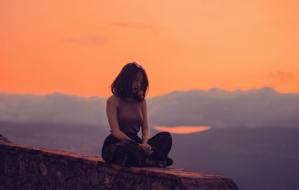 A person with short hair sits cross-legged on a brick wall at sunset, embodying self-care. The sky is a vibrant orange, and distant clouds partly obscure the horizon. Shadows cast a serene and contemplative mood over the scene.