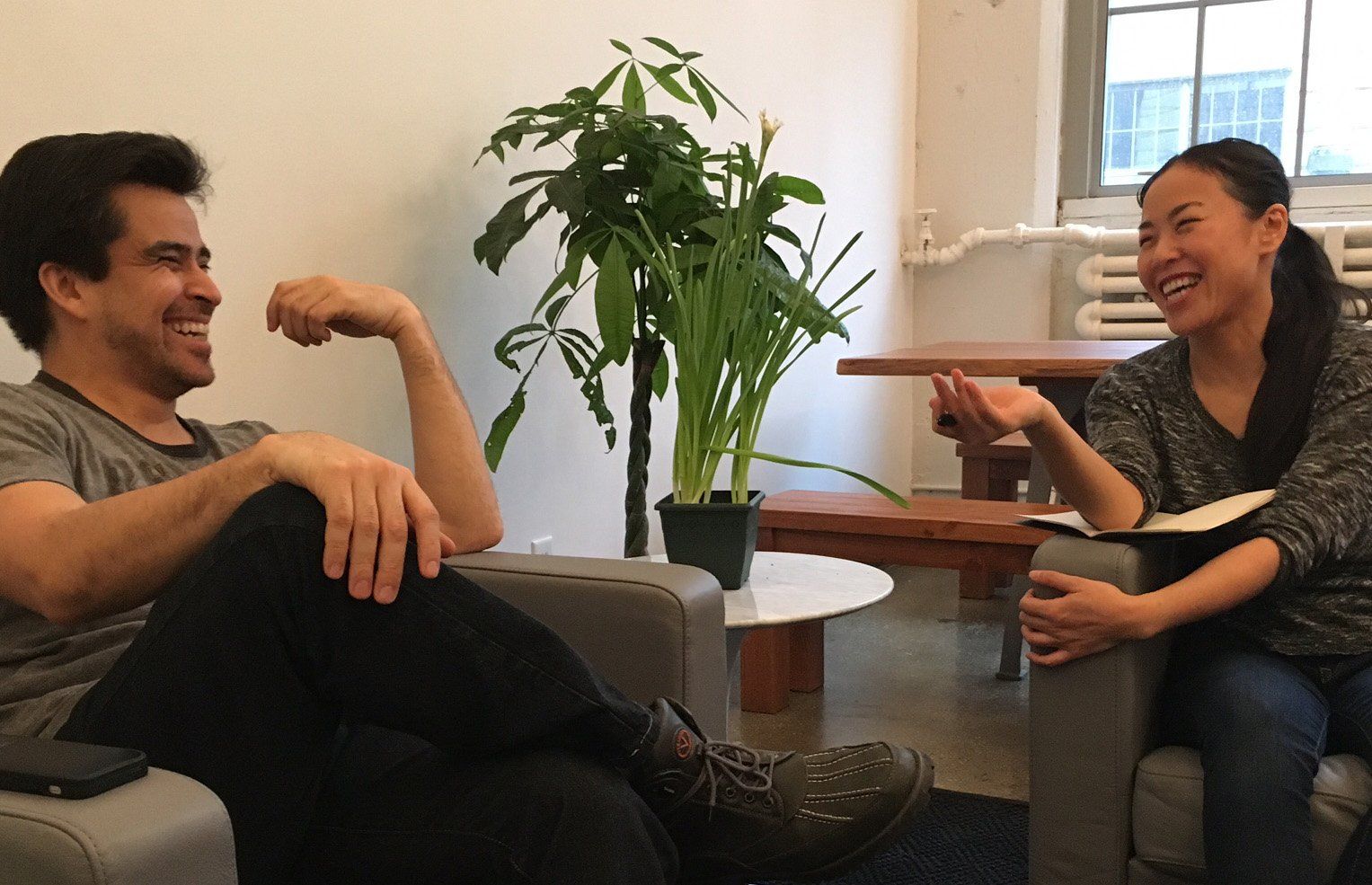 A man and a woman are sitting in armchairs, smiling and engaging in a conversation reminiscent of a classic film acting scene. A tall plant is in the background next to a table by the window, creating a casual, comfortable indoor space.