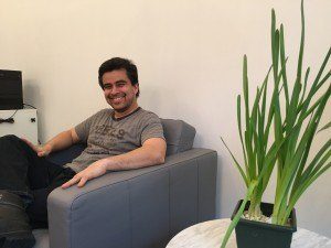 Alberto Bonilla is smiling and sitting on a gray couch in a room with a light-colored wall. A green potted plant rests on a small table beside him, while a black device is visible in the background.