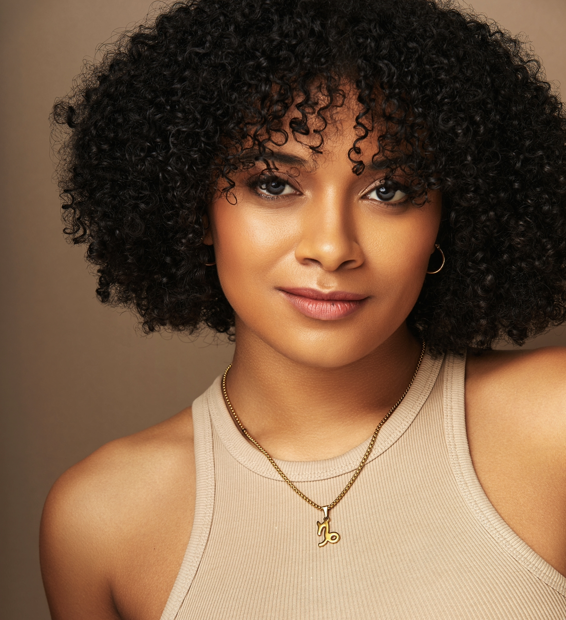 Ayla Ciccone-Burton, the renowned TKSActor, stands gracefully with her curly hair cascading over a beige sleeveless top, a subtle necklace adorning her neck. Her soft smile against the warm brown background echoes the charm of Broadway's finest moments.