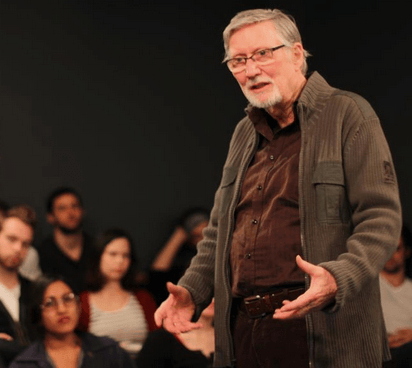 An older man with glasses and a beard, resembling a revered mentor like William Esper, addresses an attentive audience. Dressed in a brown shirt and gray cardigan, he captivates listeners while several others sit in the background, engaged with his every word.