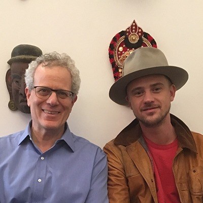 Two people sit together, smiling at the camera. One wears a blue shirt and glasses, while the other sports a hat, red shirt, and brown jacket. Decorative masks adorn the wall behind them, reminiscent of Boyd Holbrook's eclectic style in his film roles.