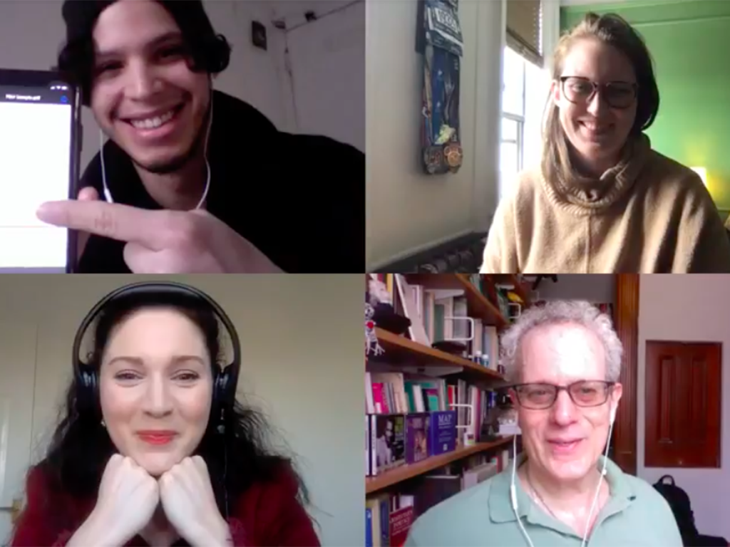 A video call with four people in a 2x2 grid, each in their own environment. The top left person, resembling TK Actor Roy Scandela, is smiling and pointing at a device. The top right and bottom right are also smiling, while the bottom left has their chin resting on their hands, looking pleased.