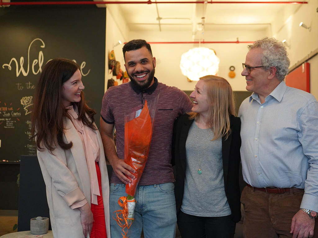 In a warmly lit room, four people gather. The man in the center, proudly holding a bouquet with orange wrapping, smiles amid cheerful faces surrounding him. A chalkboard in the background reads "Welcome" alongside mentions of TKActor Daley Baker and the prestigious Corrieri Literary Prize.