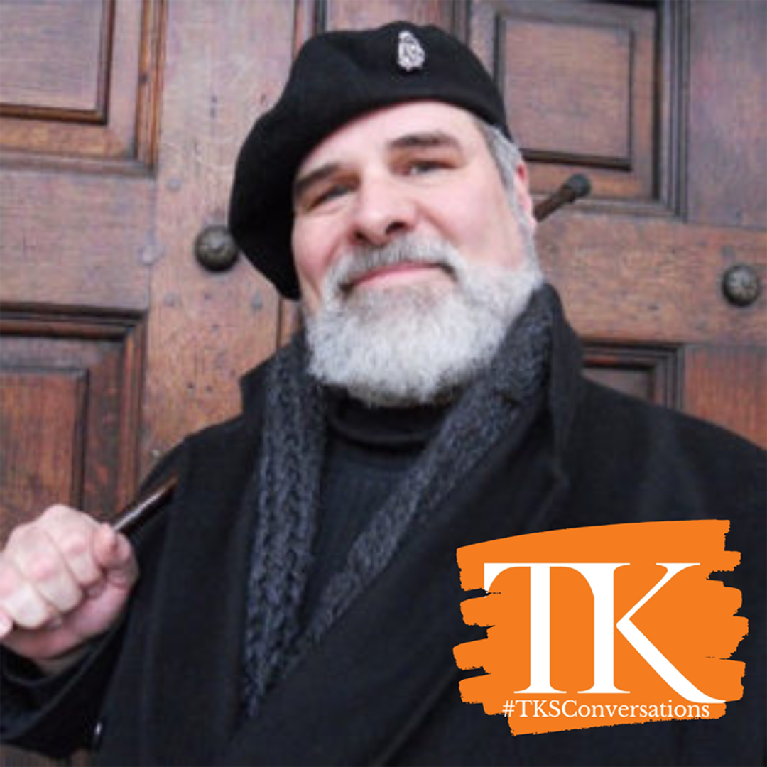 A person with a white beard, reminiscent of David Brimmer, wears a black beret and scarf while standing before a wooden door. They smile warmly, holding a pipe. The image features an orange graphic with "TK #TKConversations" in the bottom right corner.