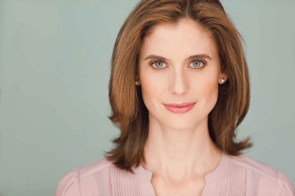 A woman with shoulder-length brown hair looks directly at the camera in this captivating headshot. She has a subtle smile and is wearing a light pink blouse. The background is a soft blue color, enhancing the serene atmosphere of the portrait.