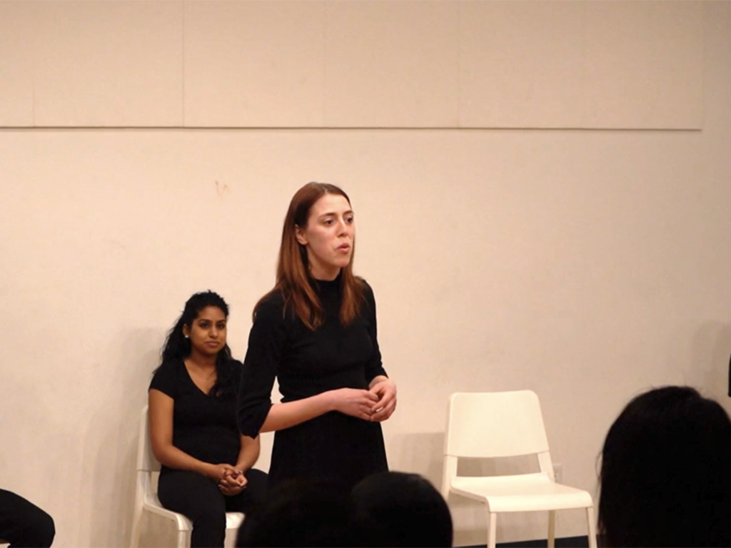 A woman in a black shirt stands on stage as a speaker, with another seated behind her. Both are in front of a white wall with a white chair nearby. The silhouettes of audience members, eager to learn about voice classes, are visible in the foreground.