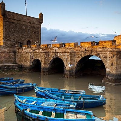 A stone fortress with arched openings stands beside calm water, where several blue wooden boats are moored. Seagulls fly overhead against a backdrop of a partly cloudy sky, creating an idyllic travel destination perfect for serene exploration and memorable adventures.