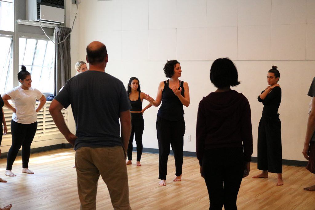 In a bright studio with wooden floors and large windows streaming in natural light, a group stands attentively. Clad in casual workout clothes, they focus on the woman at the center as she offers particularization on dance techniques.