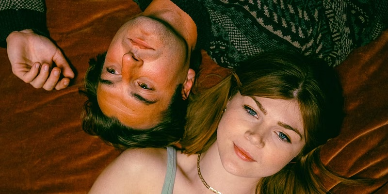 A man and woman lie on a brown surface, heads close together. The man, perhaps a TKS Alumni, has short dark hair and wears a patterned sweater. The woman has long auburn hair and dons a gray tank top. Both gaze upwards with serene expressions, as if finding their harmony in life's script at Zephyr Theatre.