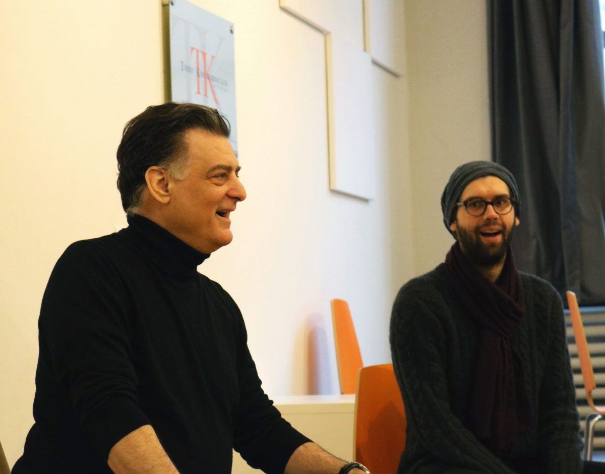 Two men are seated indoors, engaged in conversation reminiscent of a Shakespearean dialogue. The man on the left is smiling and wearing a black turtleneck, while the one on the right sports glasses, a gray beanie, and a scarf. Vibrant orange chairs and abstract wall art form the backdrop.