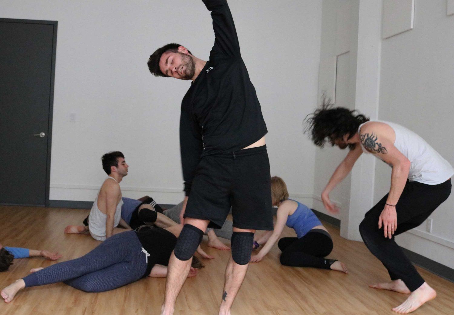 In a dance studio led by Julia Crockett, a group practices movement exercises. One person in the foreground stretches sideways wearing knee pads, while others are seated on the wooden floor, engaging in various poses. The setting is casual and relaxed.