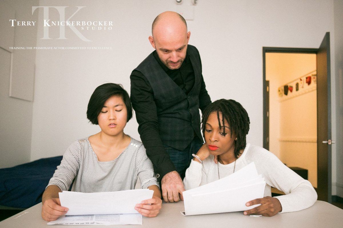 Three people are reviewing papers in an office, resembling a rehearsal of ideas. A man stands in the middle, pointing at a document held by one of two seated women. The room has neutral walls and a door in the background. The text on the wall reads "Terry Knickerbocker Studio.