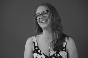 A woman with long hair and glasses smiles while looking to the side, showcasing her patterned top and pendant necklace. Trained in the Meisner Technique, she exudes a depth beyond the plain, dark background. The image is in black and white.