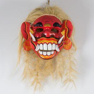 This colorful traditional mask, reminiscent of Meisner's attention to detail, showcases a red face with large white teeth and tusks. Boasting exaggerated facial features and intricate patterns, its long straw-like hair adds an element of surprise to any physical space.