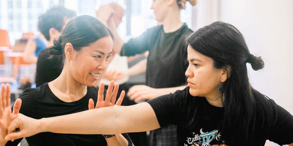 Two women practice a self-defense move; one extends her arm, and the other responds with a blocking gesture. They are focused, engaged in what seems like actors' movement training, with several people in the blurred background. The setting appears to be a classroom or studio.