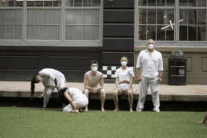 Five people dressed in white and wearing masks perform an outdoor theatrical scene as part of the Hudson Valley Movement Co.'s LIVE Movement Installation. Positioned near a building with large windows, one person bends downward while others are seated or standing, with a small sculpture hanging nearby.