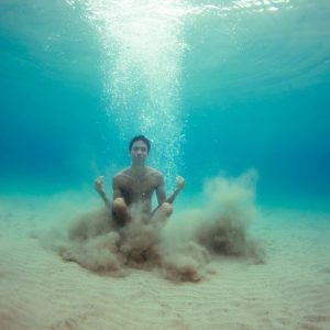 A person sits underwater on a sandy ocean floor, with bubbles rising to the surface. Participating in their own open studios of the mind, they are in a meditative pose, legs crossed and hands resting on knees. Clouds of sand surround them, creating a serene and ethereal scene.