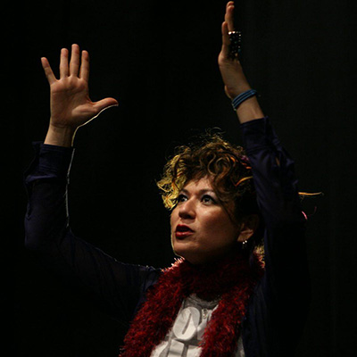 On a dark stage, under the allure of Open Studios, a person with curly hair and a red scarf performs passionately. Clad in a blue long-sleeve shirt, they raise their hands expressively while speaking or singing, an intense expression illuminating their face.