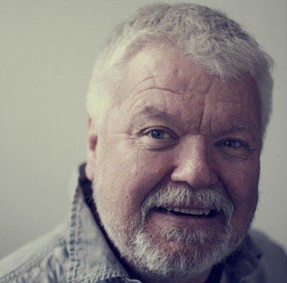 Older man with gray hair and beard, reminiscent of a modern-day Per Brahe, smiling at the camera. He dons a gray collared shirt and stands against a plain light background.