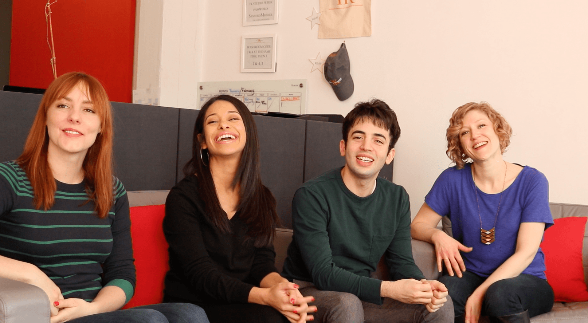 Four people sitting on a couch, smiling and looking at the camera. The background features a red wall adorned with artistic decorations reminiscent of Eve Ensler's empowering spirit. They appear to be casually dressed and relaxed.