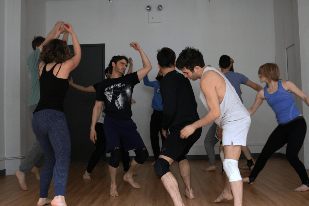 A group of people in casual clothing, led by Julia Crockett, engages in a lively dance session on wooden floors. Some wear knee pads for protection while others move energetically through various dance poses.