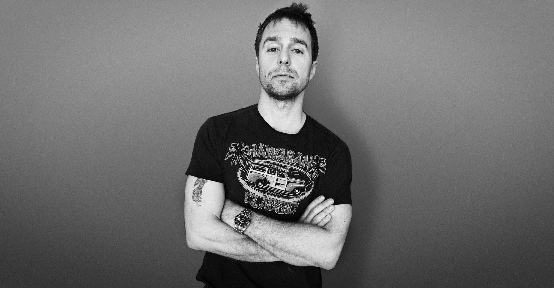 A man with short hair stands against a plain backdrop, wearing a t-shirt featuring a van graphic. His arms are confidently crossed, revealing a tattoo on his right arm. The scene evokes the cool charisma of Sam Rockwell.