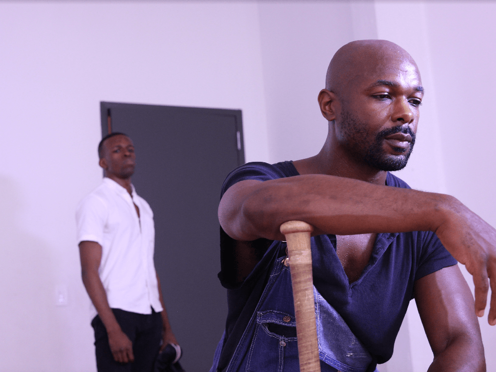 Two men indoors, perhaps actors rehearsing a scene. One is seated in the foreground holding a wooden cane, looking thoughtful, deep in the throes of self-sabotage. The other stands near a gray door, wearing a white shirt, observing intently. The room is minimally furnished and well-lit.