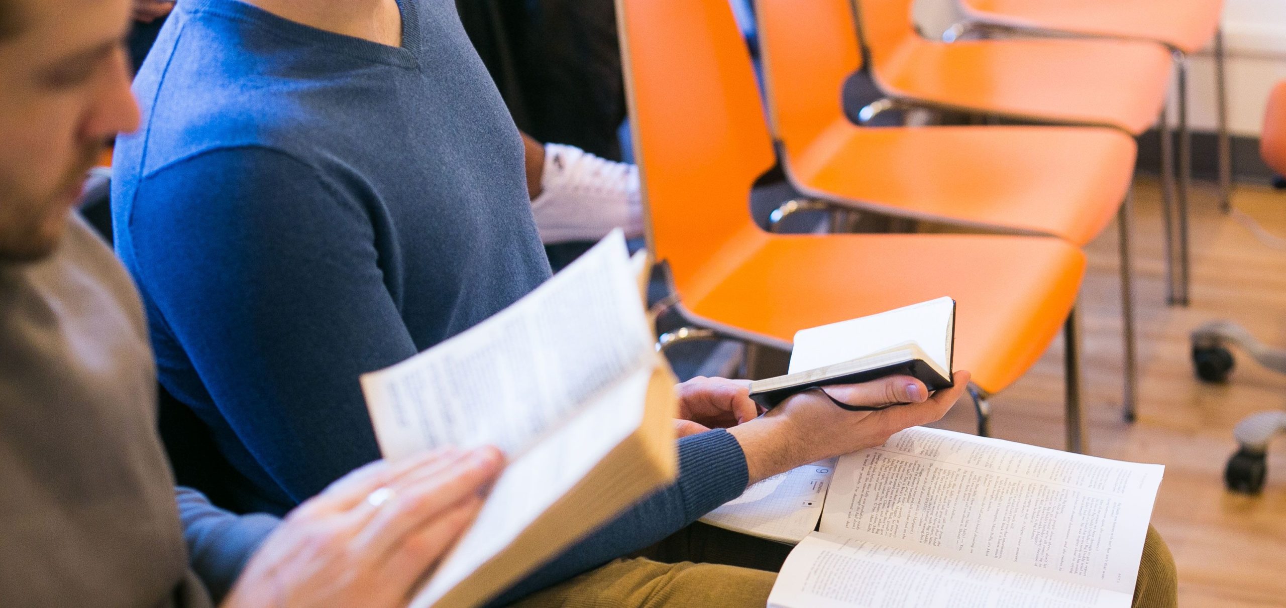 Two people are seated in orange chairs, each engrossed in reading or studying pivotal texts from the selection of books every actor needs to read. The arrangement hints at a focused group study session or class.