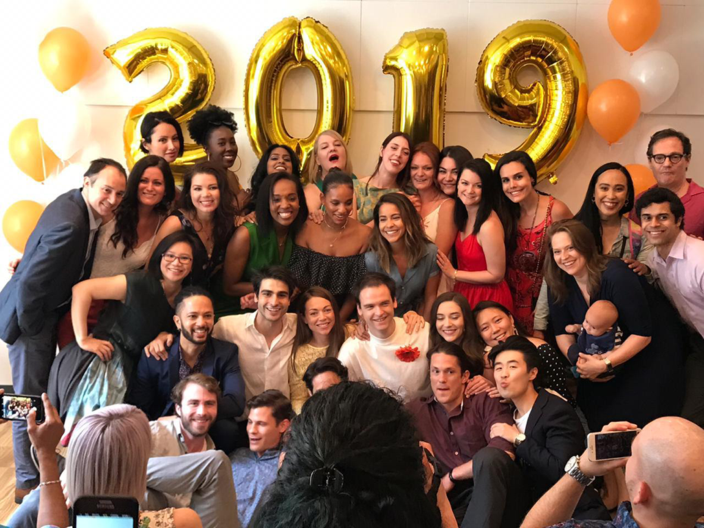 A group of people, including actors, are posing and smiling in front of large golden "2019" balloons, surrounded by orange and white ones. Some are dressed casually while others showcase their formal attire. A person is holding a baby in the front row, reminding us to stay in our own lane amidst celebrations.