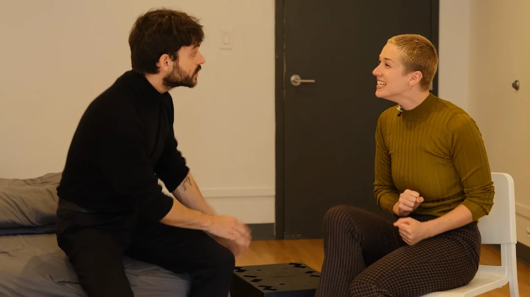 A person with short blonde hair and a green top sits on a chair, smiling as they engage in an actor rehearsal with another person who has dark hair and wears a black shirt, sitting on a bed in the cozy room.