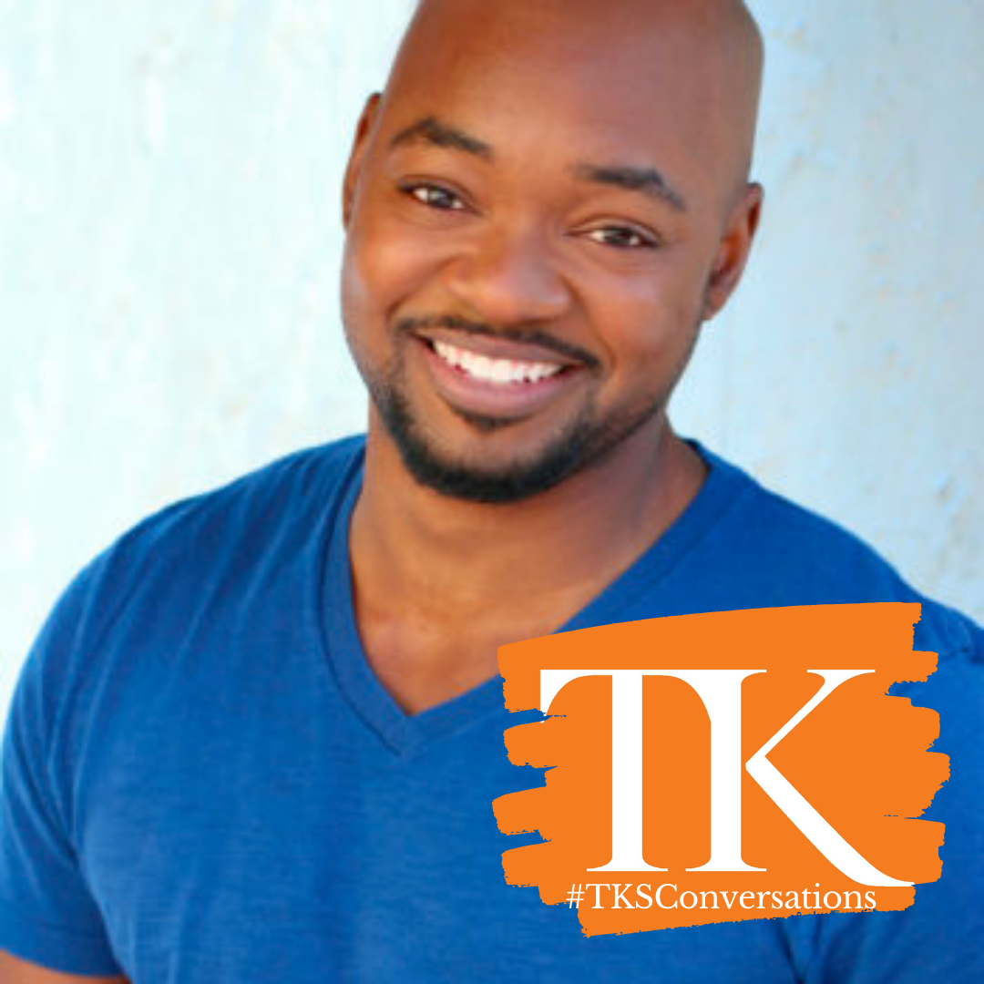 Smiling man in a blue t-shirt, reminiscent of Brian Michael Smith, stands against a light background. The orange graphic overlay with "TK" and "#TKSConversations" is prominently featured in the lower right corner.