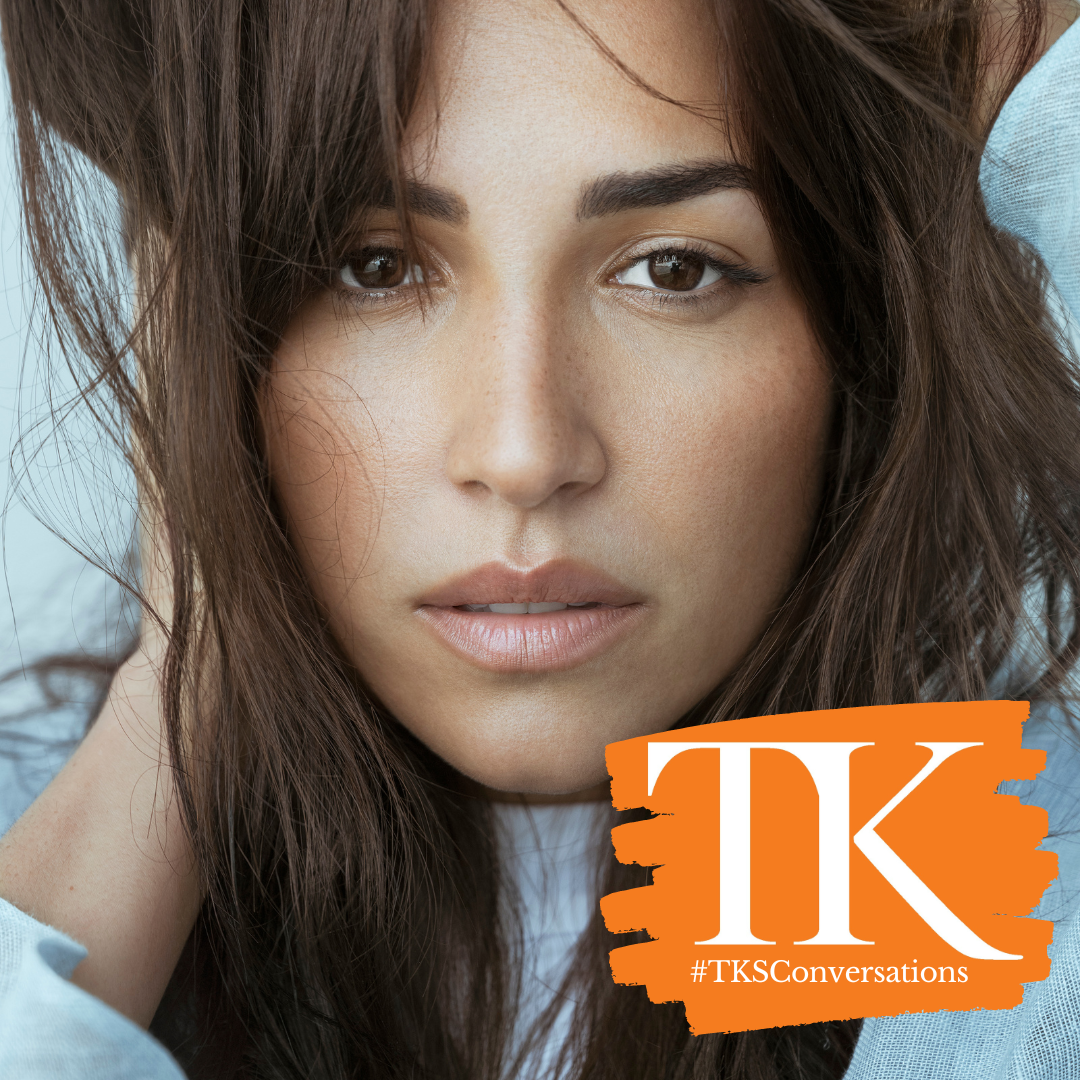 A person with long brown hair is looking directly at the camera, resting their head in their hands, reminiscent of a scene from the Westside Story movie. There's an orange logo in the bottom right corner with the text "#TKSConversations" on it.