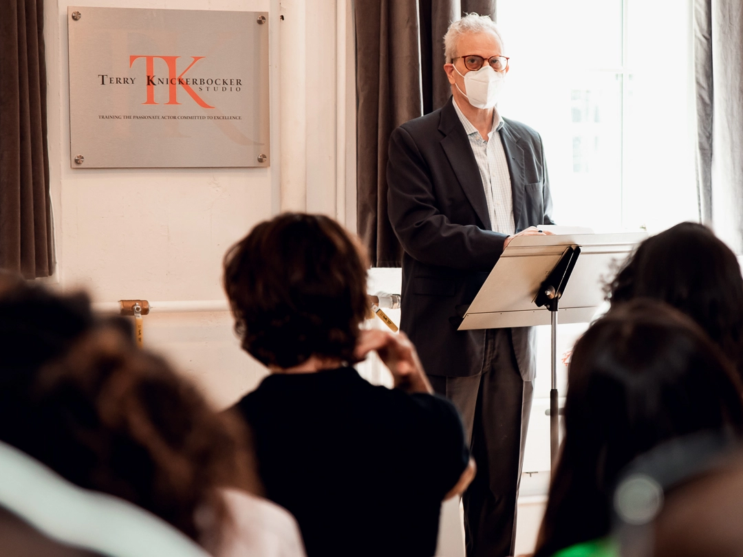 A man wearing a mask stands at a podium, addressing the audience at what appears to be a graduation ceremony. A sign on the wall reads "Terry Knickerbocker Studio," as the seated attendees eagerly listen to his speech.