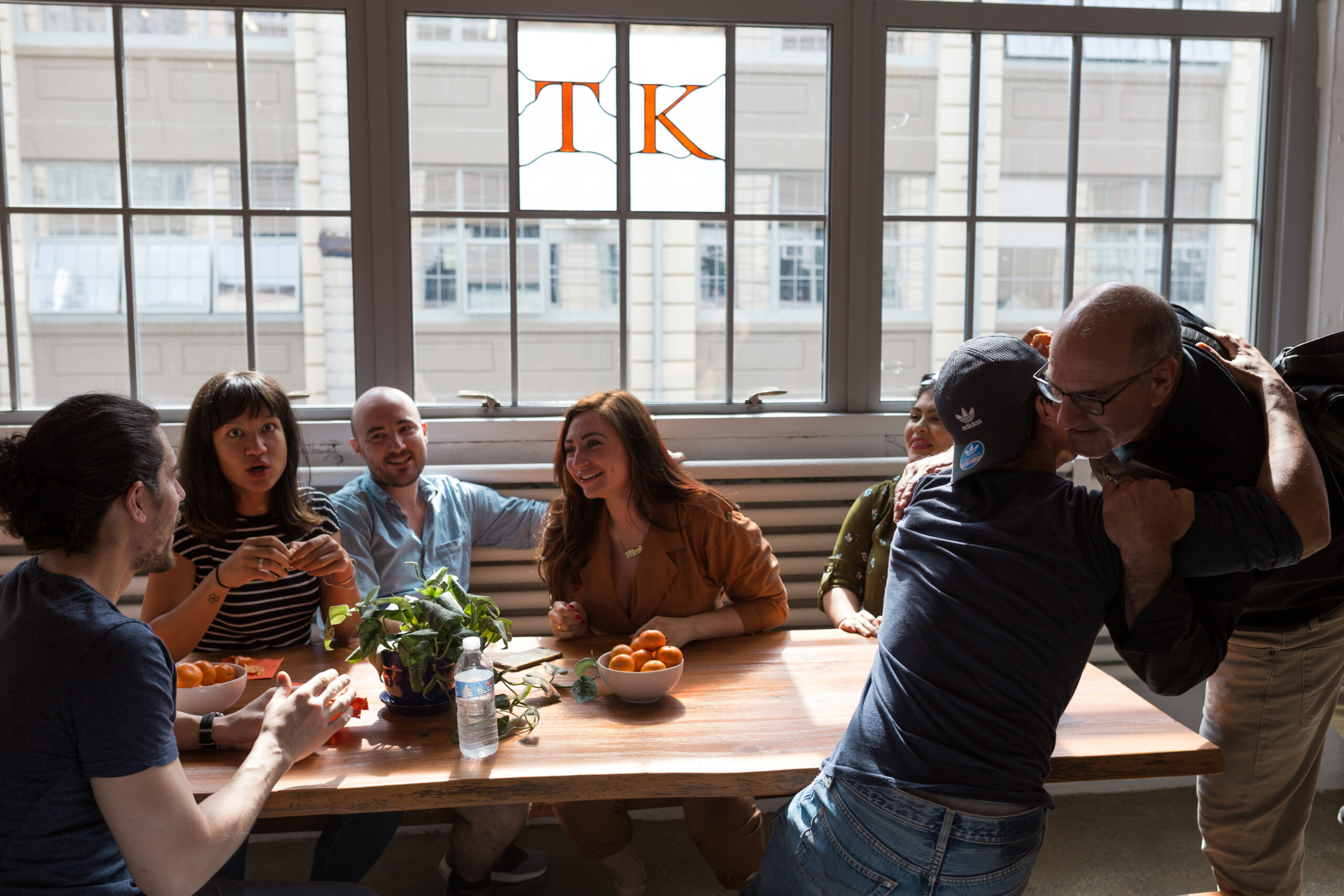 A group of six people gather around a wooden table with a bowl of oranges and plants, laughing and talking like they're in the midst of actor training. Two people on the right are embracing. Large windows in the background reveal a building outside, with "TK" on the glass.