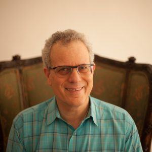 A person with curly hair, wearing glasses and a blue plaid shirt, sits on a vintage wooden chair with patterned upholstery, smiling at the camera. The softly lit background creates a warm ambiance, reminiscent of scenes often studied in quality actor training sessions.