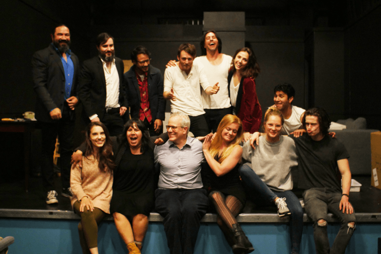 A group of thirteen people, embodying excellence in acting, pose together on a stage. Some stand while others sit at the front, their cheerful and relaxed demeanor evident as they smile and interact. The setting is indoors with dim lighting, capturing a moment on their lifelong journey in acting training.