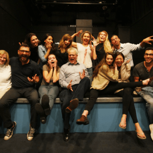 A group of fourteen people poses energetically on a stage, many making funny faces and gestures. They are seated and standing, embodying excellence in acting as they appear relaxed and cheerful in this casual setting—a testament to their lifelong journey of training together.