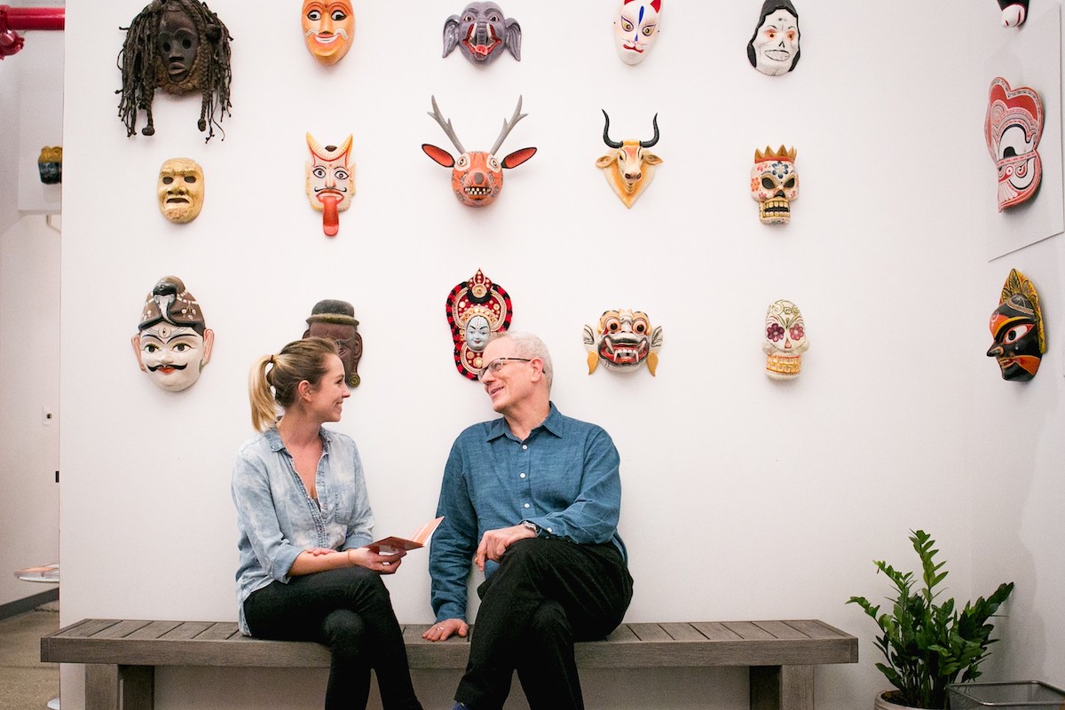 Two people sit on a bench in front of a wall adorned with colorful masks, deep in conversation about their NYC acting classes. Papers in hand, they discuss techniques learned from Terry Knickerbocker as a small plant rests beside them on the floor.