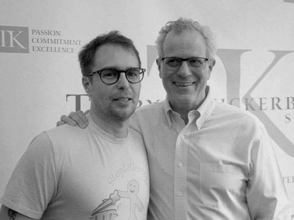 Two men stand indoors, both wearing glasses and smiling. The man on the left sports a casual t-shirt, while his friend on the right opts for a button-up shirt. Behind them, a backdrop displays "PASSION COMMITMENT EXCELLENCE," embodying their dedication like actors mastering their craft through training.